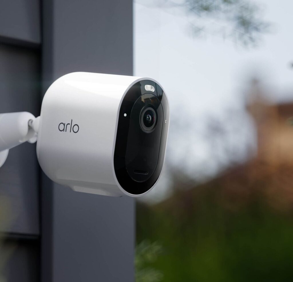 A white Arlo Pro 5S 2K Spotlight Camera is mounted on an outdoor wall, facing slightly to the right with greenery and an out-of-focus building in the background.